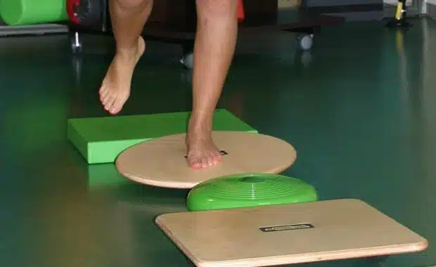 Una persona è in equilibrio su un piede su una balance board circolare, che mostra la rieducazione propriocettiva Roma Prati presso Fisioprati. Altri attrezzi per l'equilibrio appaiono sul pavimento, suggerendo una palestra indoor o un centro di riabilitazione.
