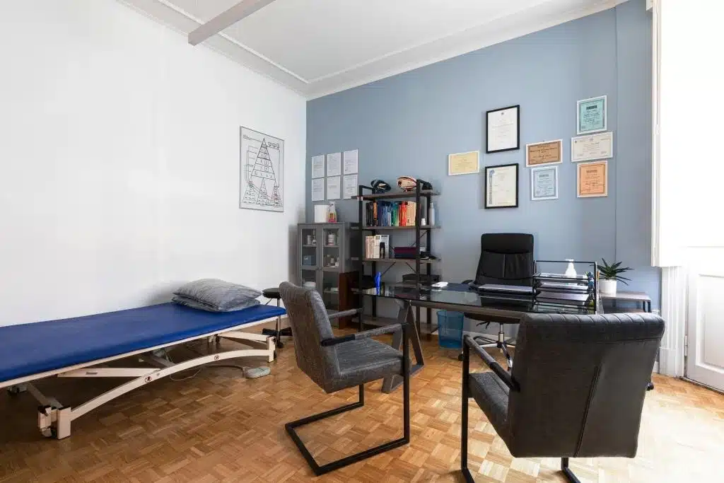 Uno studio medico con una scrivania, due sedie, un lettino per le visite, librerie e certificati incorniciati alla parete presso lo studio Fisioprati Roma.