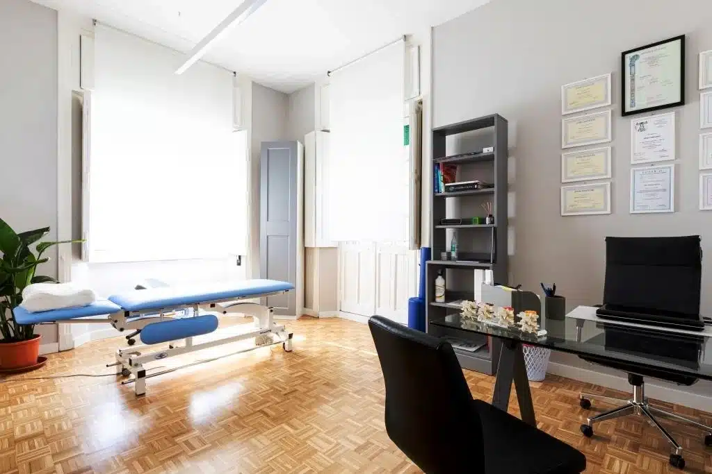 Un luminoso ufficio in Via Giovanni Bettolo 6, Roma, è dotato di un lettino per visite mediche, una scrivania, una libreria, certificati alla parete, una pianta in vaso e ampie finestre con persiane bianche.