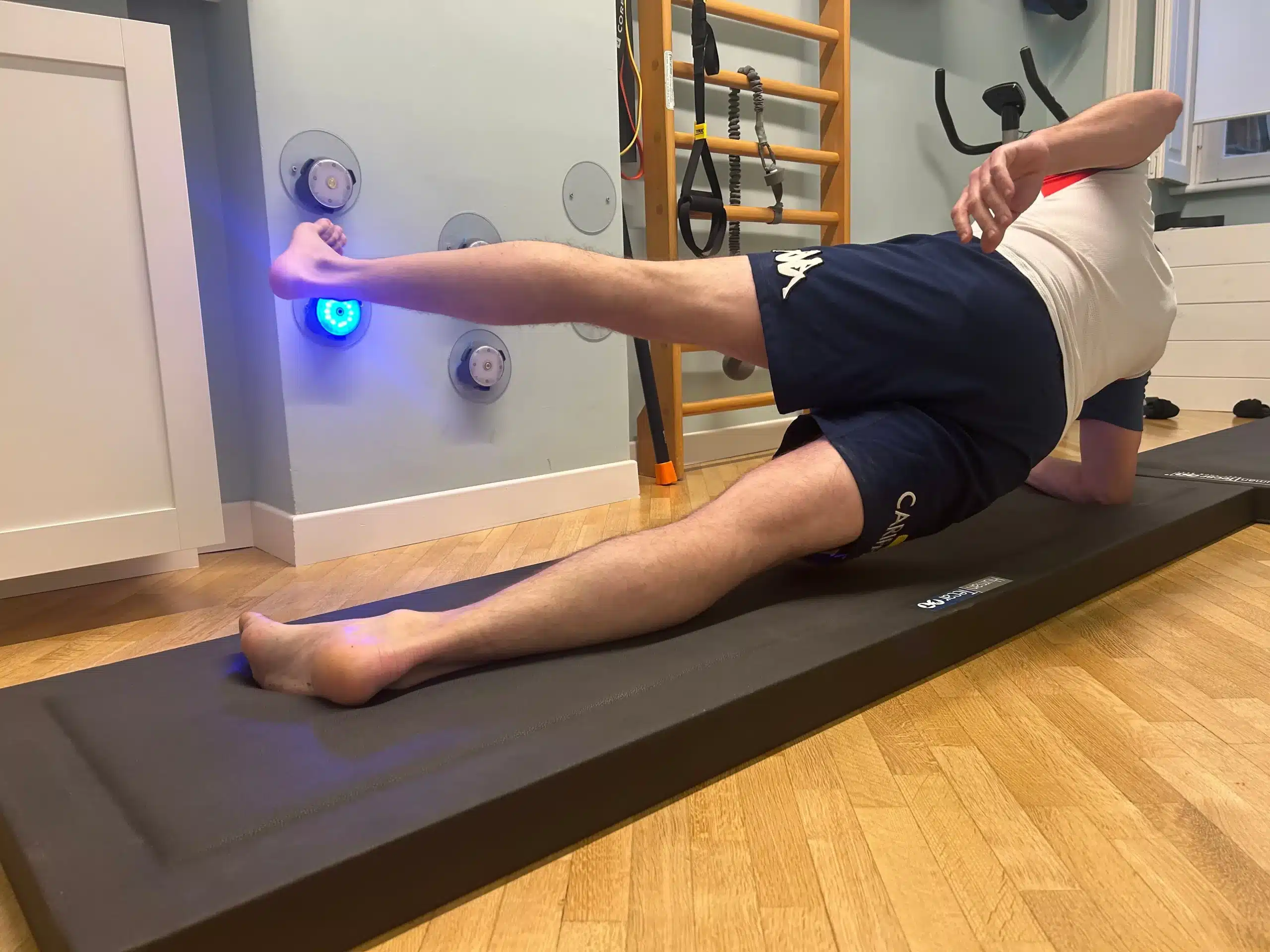 Una persona esegue un esercizio di sollevamento delle gambe in posizione laterale su un tappetino accanto a un'attrezzatura per esercizi montata a parete in una stanza con pavimento in legno, partecipando al programma Ritorno allo Sport: Riabilitazione NMRtS presso Fisioprati Roma.