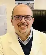 Un uomo con occhiali e camice bianco, identificato come Dott. Di Paola Berardino, sorride mentre è seduto in un ufficio con documenti e una foto sullo sfondo.