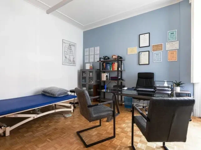 Uno studio medico con una scrivania, due sedie, un lettino per le visite, librerie e certificati incorniciati alla parete presso lo studio Fisioprati Roma.