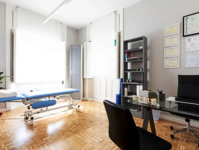 Un luminoso ufficio in Via Giovanni Bettolo 6, Roma, è dotato di un lettino per visite mediche, una scrivania, una libreria, certificati alla parete, una pianta in vaso e ampie finestre con persiane bianche.
