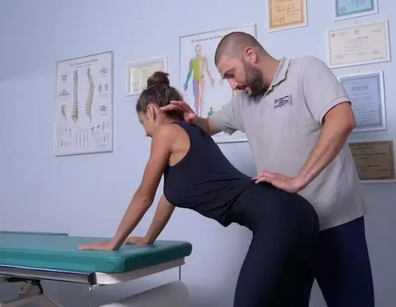 Un fisioterapista presso FisioPrati di Roma guida una donna in abito nero in un esercizio per la schiena per la riabilitazione motoria, con certificati e tabelle anatomiche appese alla parete alle loro spalle.